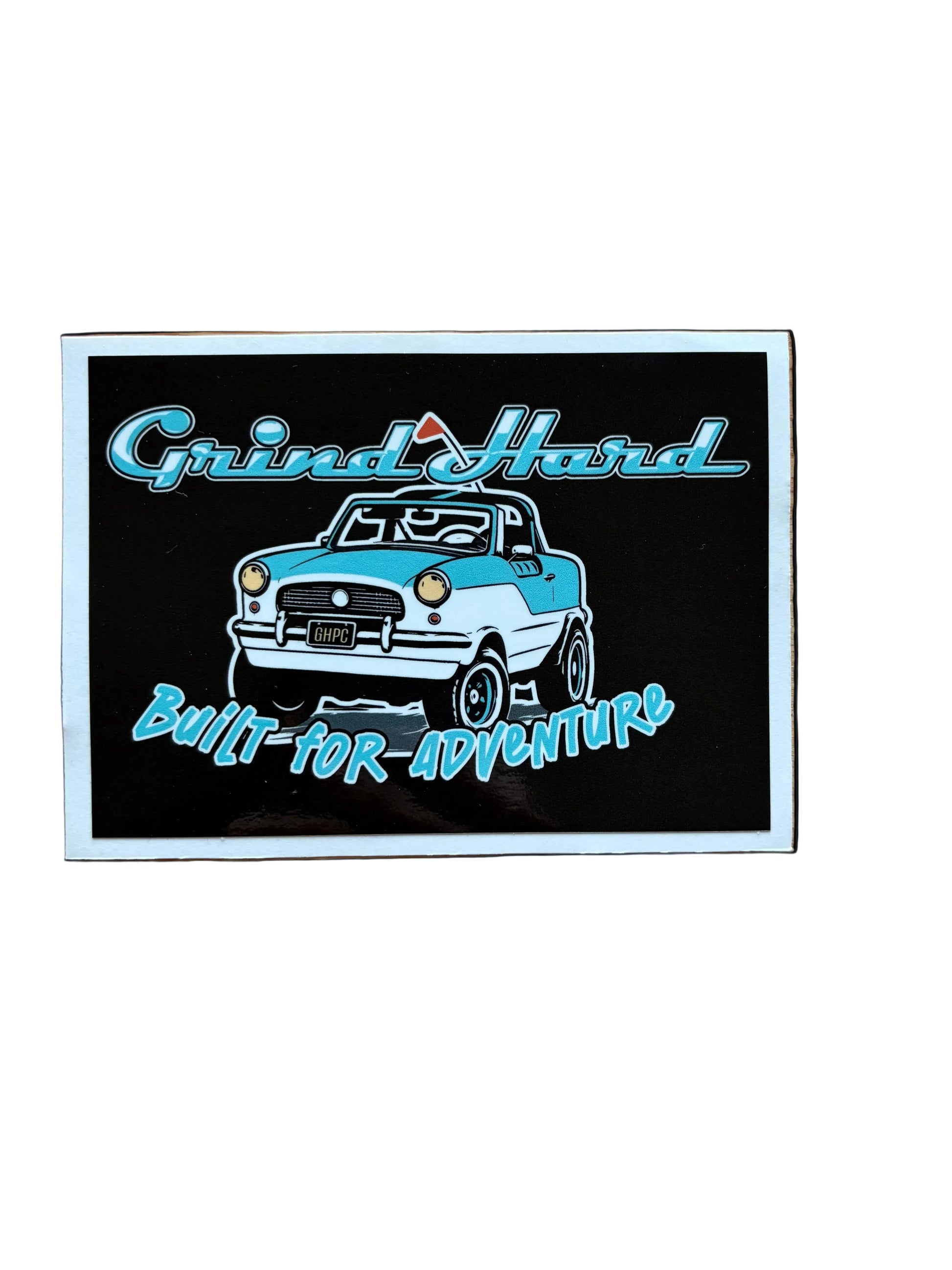 A rectangular sticker with a white border lies on a light brown wooden surface. The sticker features a black background with a graphic of a light blue and white vintage car, stylized text reading 'Grind Hard' at the top and 'Built for Adventure' at the bottom, and a small mountain icon above the car.