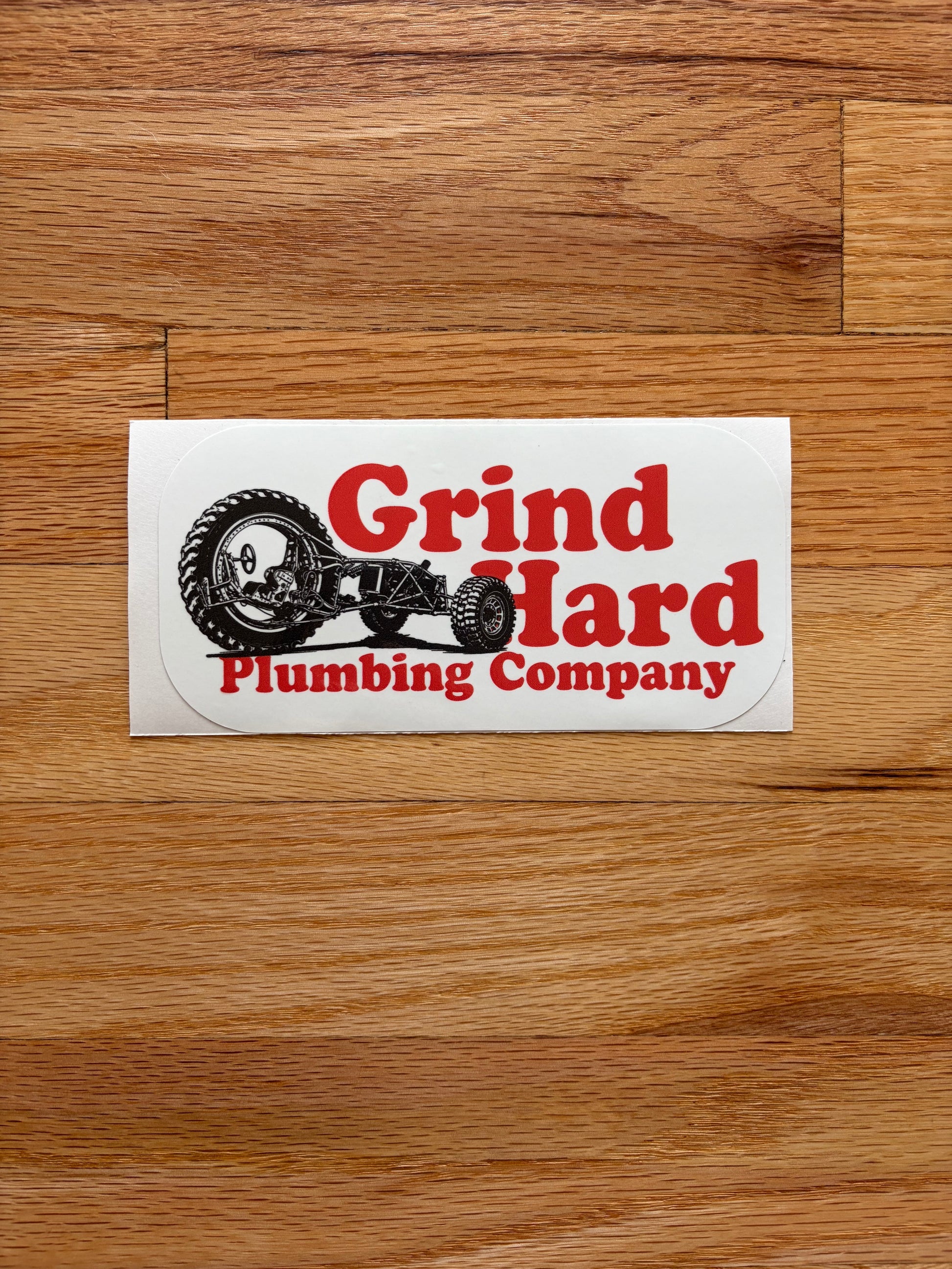 A rectangular white sticker with red and black text reading 'Grind Hard Plumbing Company' and a black tractor illustration, placed on a brown wood surface.
