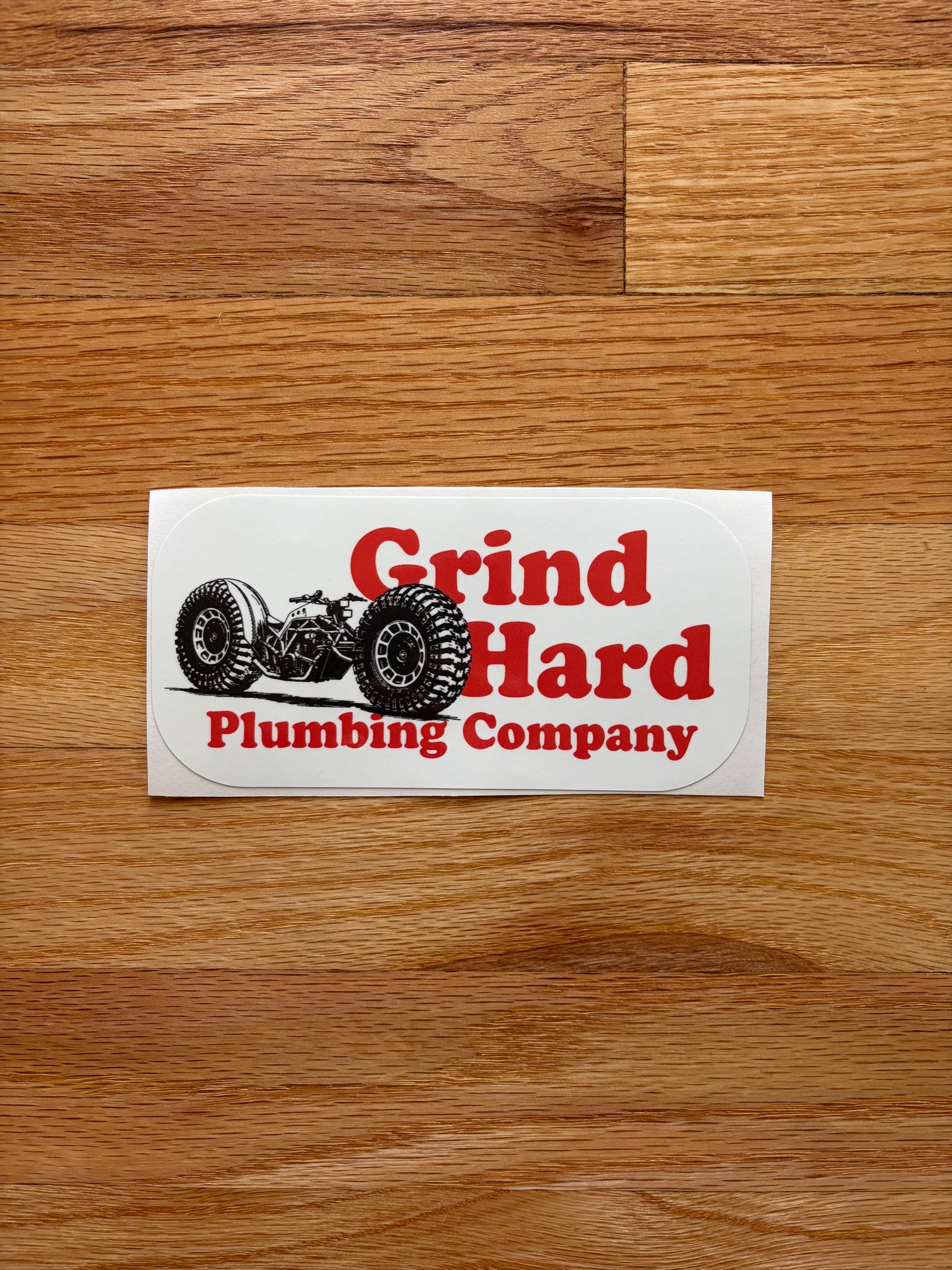 A rectangular white sticker with a red and black graphic of a tractor and the text 'Grind Hard Plumbing Company' lies on a brown wooden floor.
