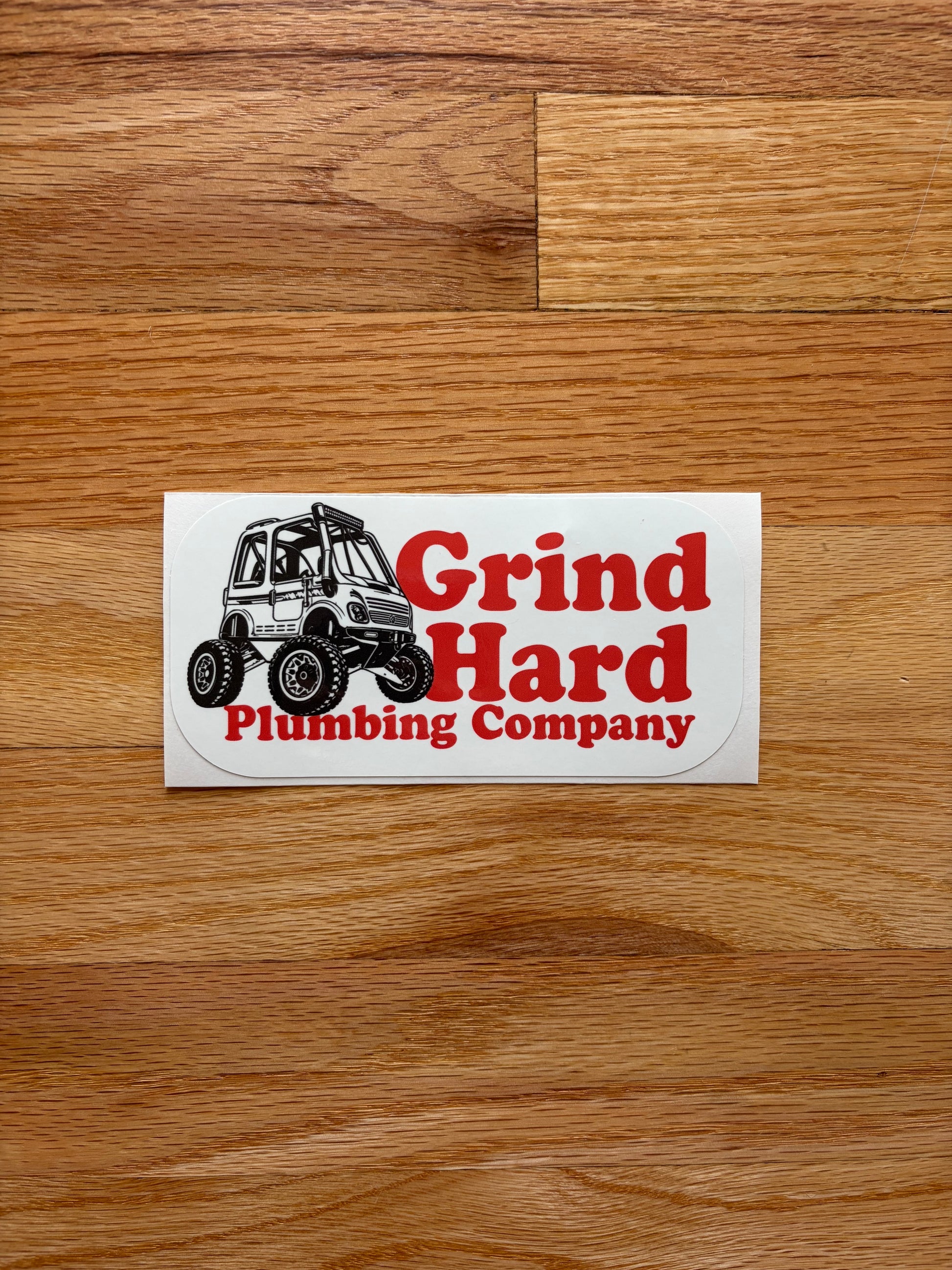 A rectangular white sticker with red and black text reading 'Grind Hard Plumbing Company' and an illustration of an off-road vehicle, placed on a brown wood plank surface.