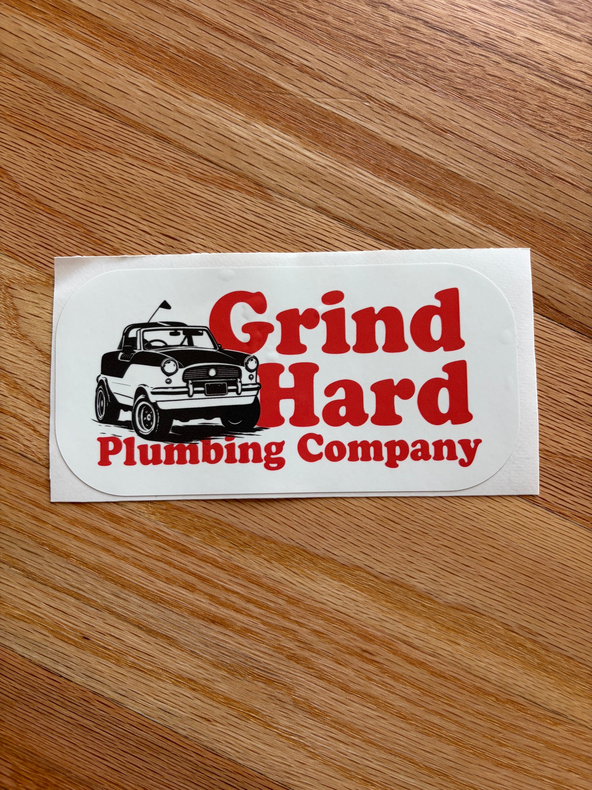 A white die-cut sticker with the red text 'Grind Hard Plumbing Company' and a black-and-white illustration of a car, placed on a wooden surface.